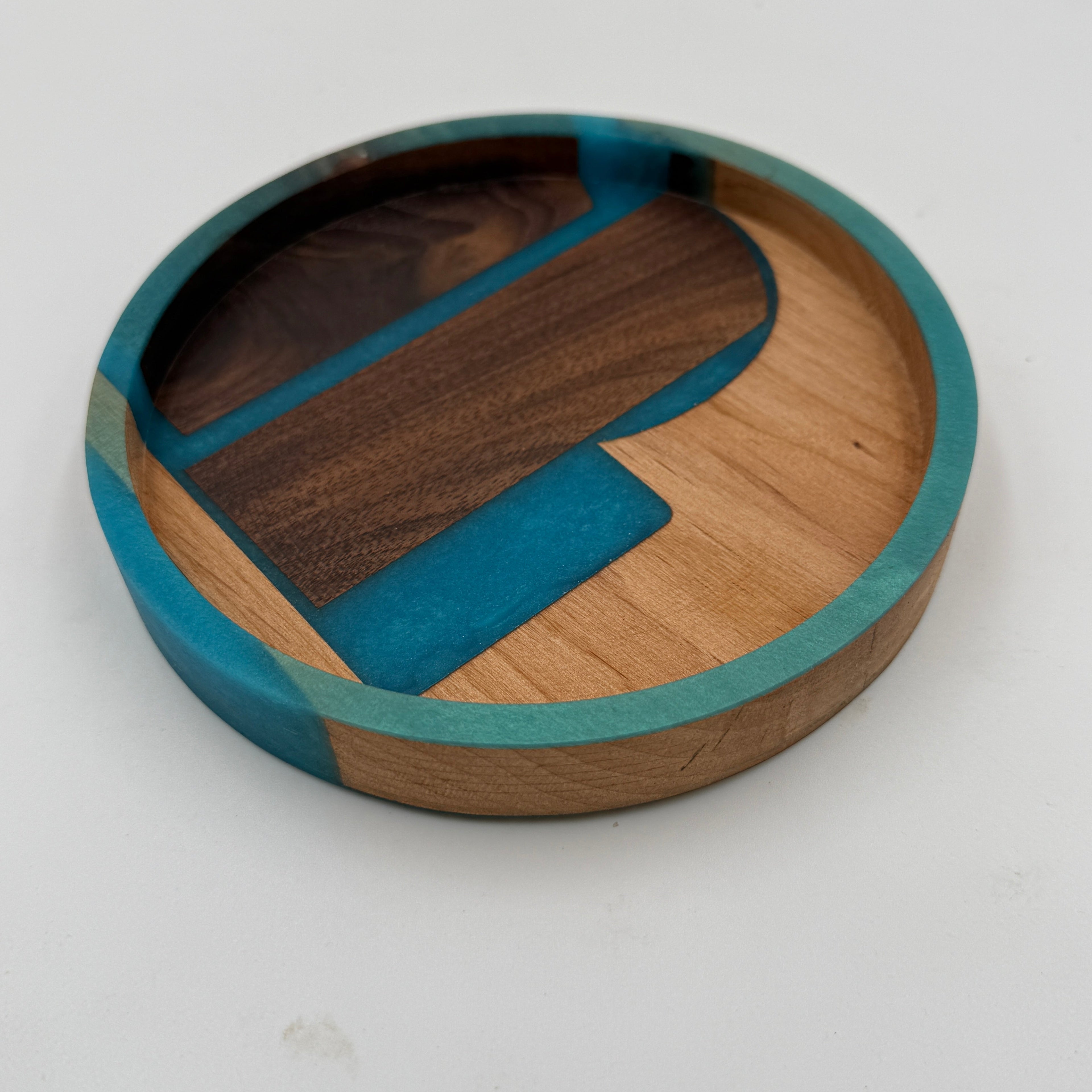 A round wooden tray with a colorful inlay design, featuring a teal outer rim, a dark brown wood section, a light natural wood section, and a bright teal accent piece, set against a plain white background.